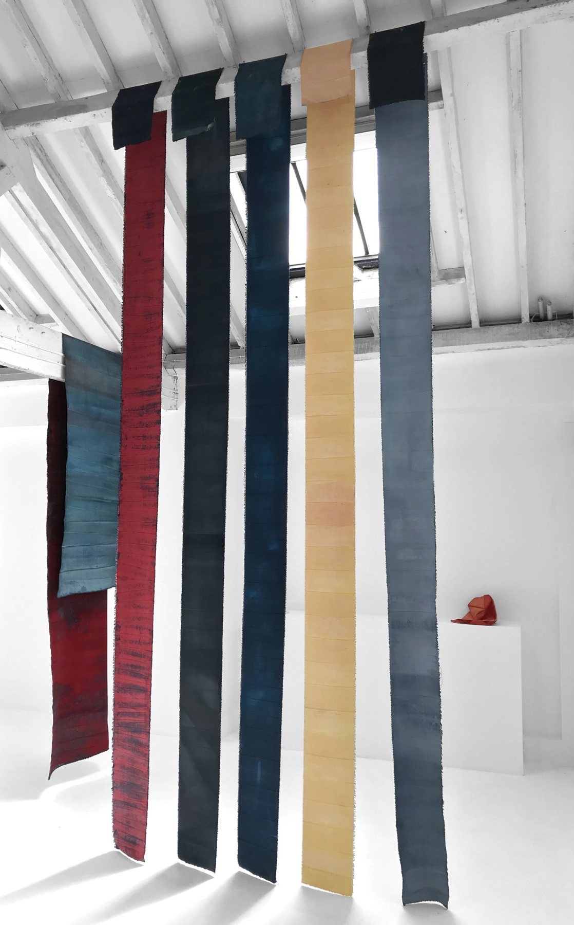 Long unstretched canvas drops in red, navy, grey and ochre hanging from ceiling to floor in a white gallery. Eutopia, 2019 by Emma Hurst.