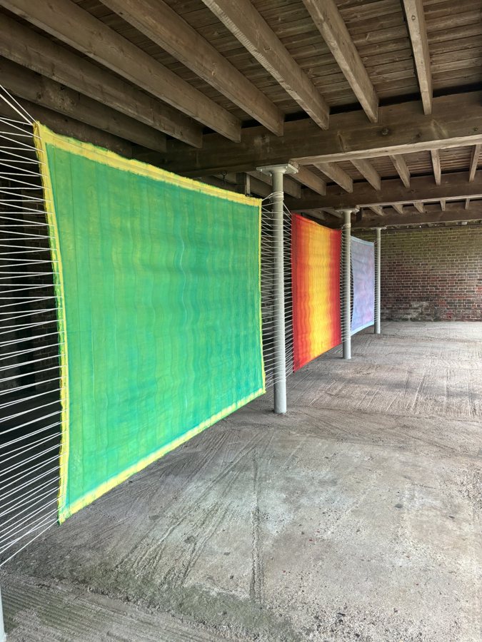 Three canvas panels in green, turquoise and orange-red stretched between the pillars of a barn undercroft at South Stoke. Untitled (Idyll), 2024, by Emma Hurst.