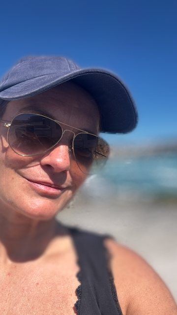 Emma on a beach in a cap and sunglasses, sea behind her